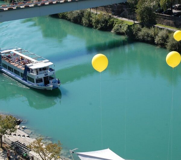 Ein Passagierboot fährt auf einem türkisfarbenen Fluss unter einer Brücke hindurch, in dessen Nähe mehrere gelbe Luftballons schwimmen und Bäume das Ufer säumen.