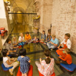 Eine Gruppe von Kindern und ein Erwachsener sitzen im Kreis auf Kissen auf einem Glasboden in einem Raum mit gewölbten Ziegelwänden.