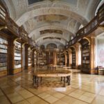 Eine prächtige, verschnörkelte Bibliothek mit hohen Holzregalen, gewölbten Deckenfresken und einem runden Holztisch in der Mitte des polierten Bodens.