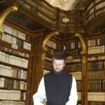 Ein Mann in einer schwarz-weißen Weste liest ein großes Buch an einem Tisch in einer kunstvollen Bibliothek mit hohen Regalen und gewölbten Decken, die mit Büchern gefüllt sind.