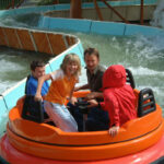 Vier Personen fahren auf einem runden orangefarbenen Floß auf einer Wasserbahn, im Hintergrund sind spritzendes Wasser und gebogene Barrieren zu sehen.