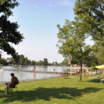 Menschen entspannen sich an einem grasbewachsenen Flussufer mit Bäumen, einige sitzen auf Stühlen, andere am Wasser; sonniges Wetter und ruhiges Wasser im Hintergrund.