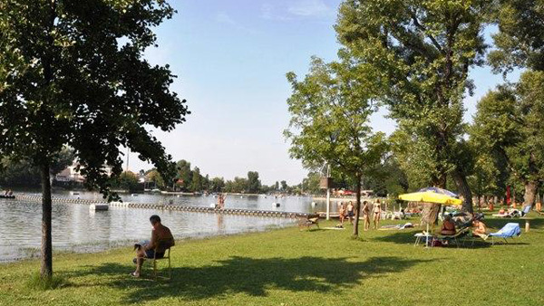 Menschen entspannen sich an einem grasbewachsenen Flussufer mit Bäumen, einige sitzen auf Stühlen, andere am Wasser; sonniges Wetter und ruhiges Wasser im Hintergrund.