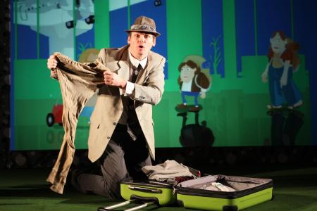 Ein Mann in Anzug und Hut kniet neben einem offenen Koffer und hält eine Hose hoch, während hinter ihm eine Reiseszene im Cartoon-Stil projiziert wird.