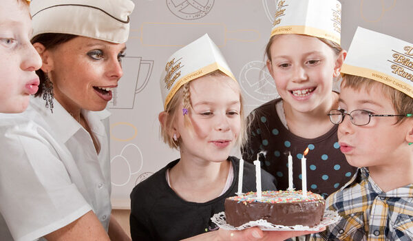 Vier Kinder und ein Erwachsener mit Papierhüten haben sich um einen kleinen Schokoladenkuchen mit brennenden Kerzen versammelt und bereiten sich darauf vor, diese gemeinsam auszublasen.