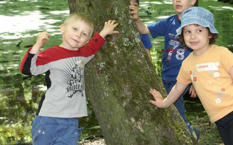 Vier junge Kinder stehen im Freien an einem Baum, wobei zwei von ihnen den Baumstamm berühren. Sie sind leger gekleidet und scheinen sich in einem Park oder Naturgebiet aufzuhalten.