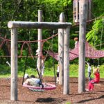 Kinder spielen auf einem Spielplatz mit Kletterseilen, Netzen und Schaukeln, umgeben von Holzpfählen und Grünpflanzen im Hintergrund.