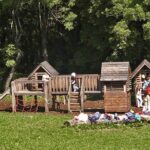 Kinder spielen auf einem hölzernen Spielplatz mit einer Rutsche und einem Kletternetz, während Taschen und Gegenstände auf dem Gras im Vordergrund verstreut sind. Der Hintergrund ist von Bäumen gesäumt.