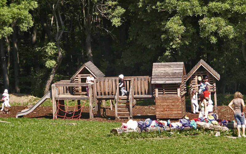 Kinder spielen auf einem hölzernen Spielplatz mit einer Rutsche und einem Kletternetz, während Taschen und Gegenstände auf dem Gras im Vordergrund verstreut sind. Der Hintergrund ist von Bäumen gesäumt.
