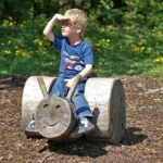 Ein kleiner Junge sitzt auf einem hölzernen Spielplatz, der die Form eines Gesichts hat, und schützt seine Augen mit der Hand vor der Sonne.