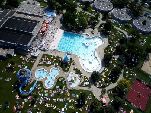 Luftaufnahme eines öffentlichen Freibadkomplexes mit Wasserrutschen, Sonnenanbetern auf der Wiese und einem nahe gelegenen Basketballplatz.