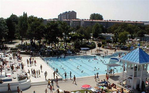 Großer Swimmingpool im Freien, umgeben von Menschen, mit Bäumen und Gebäuden im Hintergrund an einem sonnigen Tag.