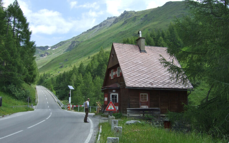 Ein Mann steht in der Nähe eines kleinen Holzhauses an einer kurvigen Bergstraße, umgeben von grünen Hügeln und Bäumen unter einem teilweise bewölkten Himmel.