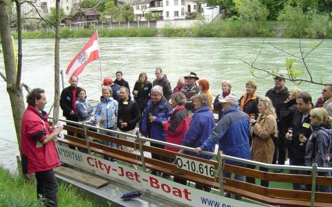 Eine Gruppe von Menschen steht auf einem angedockten Jetboot an einem Fluss und hört einem Mann in einer roten Jacke zu, der ein Mikrofon hält. Am Heck des Bootes ist eine österreichische Flagge angebracht.