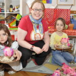 Ein Erwachsener und zwei kleine Kinder sitzen in einem farbenfrohen Raum auf dem Boden und halten rosafarbene Bastelprojekte in der Hand, umgeben von Kunstzubehör.