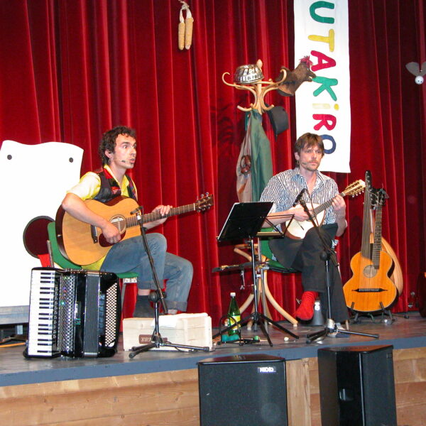 Zwei Musiker sitzen auf der Bühne und spielen Saiteninstrumente, daneben stehen ein Akkordeon und Gitarren. Im Hintergrund sind ein roter Vorhang, Requisiten und ein buntes vertikales Banner zu sehen.