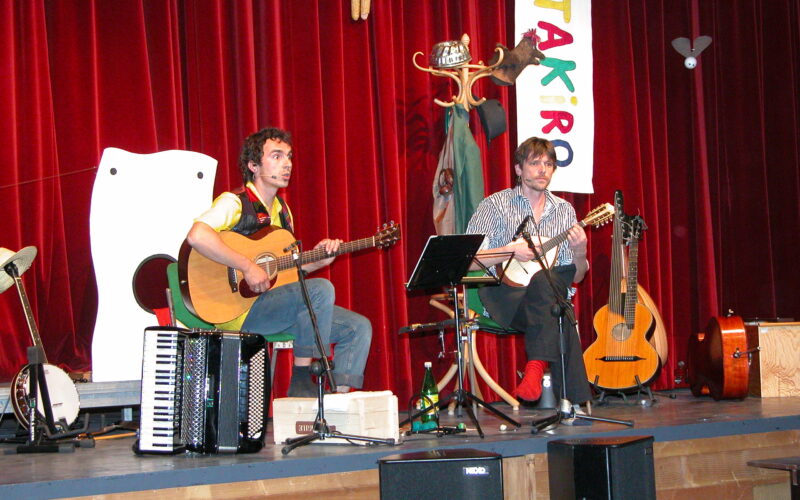 Zwei Musiker sitzen auf der Bühne und spielen Saiteninstrumente, daneben stehen ein Akkordeon und Gitarren. Im Hintergrund sind ein roter Vorhang, Requisiten und ein buntes vertikales Banner zu sehen.