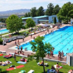 Ein großer öffentlicher Freibadkomplex, in dem die Leute schwimmen und andere sich auf der umliegenden Wiese sonnen.