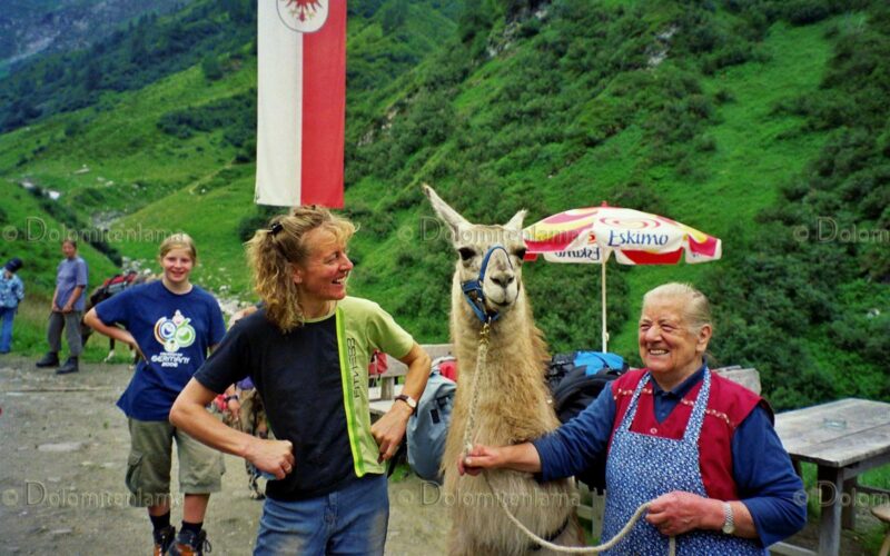 Zwei Menschen stehen lächelnd neben einem Lama auf einem Bergpfad; einer hält die Leine des Lamas. Im Hintergrund sind grüne Hügel und Flaggen zu sehen.
