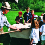 Ein Erwachsener mit Hut demonstriert einer Gruppe von Kindern, die sich im Freien um einen Tisch versammelt haben, das Backen mit einem Holzpaddel. Im Hintergrund sind Bäume und Gras zu sehen.