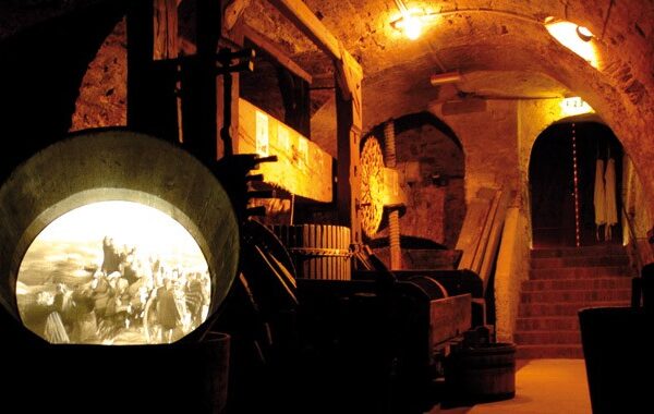 Schummerig beleuchteter Steinkeller mit gewölbten Decken, Holzbalken, Treppen, verschiedenen Werkzeugen und einer runden Auslage, die mit einem alten Foto beleuchtet ist.
