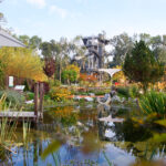Ein angelegter Garten mit einem Teich im Vordergrund, üppigen Pflanzen, einem Regenschirm und einem metallenen Aussichtsturm im Hintergrund unter einem klaren Himmel.