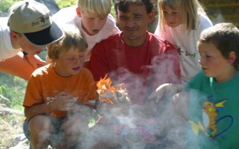 Eine Gruppe von Kindern und ein Erwachsener beobachten aufmerksam, wie ein kleines Feuer auf einem Stück Holz brennt und der Rauch um sie herum aufsteigt.