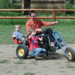 Ein Erwachsener und zwei Kinder fahren auf einem großen Dreirad mit Tretantrieb auf einem unbefestigten Weg in einer Außenanlage mit Gras und Holzzaun.