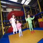 Drei Kinder springen auf einem gelben Trampolin in einem Indoor-Spielplatz mit Netzwänden und einer roten Rutsche im Hintergrund.