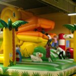 Die Kinder spielen auf einer großen, farbenfrohen, aufblasbaren Dschungel-Hüpfburg mit Palmen-Dekoration und einer orangefarbenen Rutsche in einem geräumigen Innenbereich.