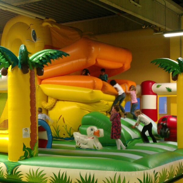 Die Kinder spielen auf einer großen, farbenfrohen, aufblasbaren Dschungel-Hüpfburg mit Palmen-Dekoration und einer orangefarbenen Rutsche in einem geräumigen Innenbereich.