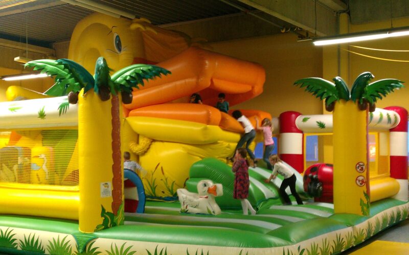 Die Kinder spielen auf einer großen, farbenfrohen, aufblasbaren Dschungel-Hüpfburg mit Palmen-Dekoration und einer orangefarbenen Rutsche in einem geräumigen Innenbereich.