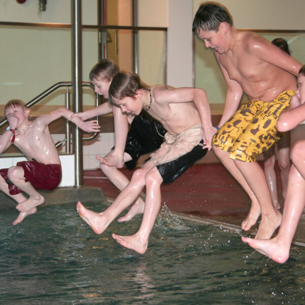 Vier Jungen in Badehosen springen gemeinsam in ein Hallenbad, mitten in der Luft, und das Wasser spritzt um sie herum.