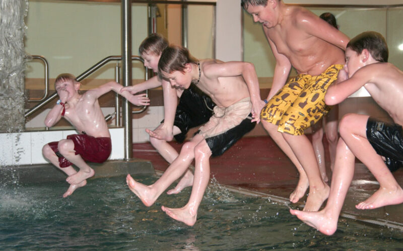 Vier Jungen in Badehosen springen gemeinsam in ein Hallenbad, mitten in der Luft, und das Wasser spritzt um sie herum.