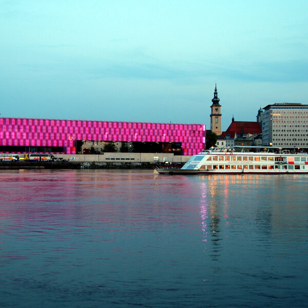 Ein modernes Flussschiff fährt an einem hell erleuchteten rosa Gebäude und einer Stadtlandschaft vorbei, die sich in der Abenddämmerung im ruhigen Wasser spiegelt.