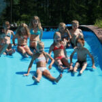 Eine Gruppe von Kindern in Badeanzügen sitzt an einem sonnigen Tag im Freien, umgeben von Bäumen und Grün, am Rande eines Pools mit Wasser.