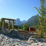 Ein aus Stein gebauter Wasserspielplatz mit Holzstrukturen, einem kleinen roten Gebäude, Bäumen und Bergen im Hintergrund unter einem klaren Himmel.