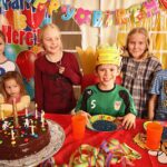 Sechs Kinder lächeln um einen Geburtstagstisch mit einem Schokoladenkuchen, Kerzen, bunten Tellern, Tassen und einem "Happy Birthday"-Banner. Ein Kind trägt eine Papierkrone.