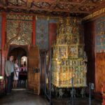 Zwei Personen stehen in einem reich verzierten Raum mit kunstvollen Goldverzierungen, Holzwänden und einem großen, dekorativen Keramikofen.