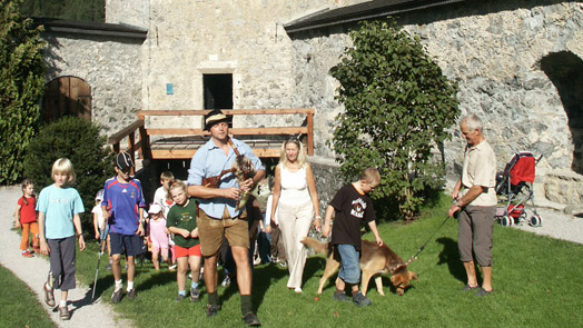 Eine Gruppe von Erwachsenen und Kindern, einige mit Ziegen an der Leine, spaziert an einem sonnigen Tag auf einem grasbewachsenen Weg in der Nähe eines Steinhauses.