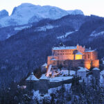 Ein großes, in der Abenddämmerung beleuchtetes Schloss steht auf einem verschneiten, bewaldeten Hügel, mit hohen, schneebedeckten Bergen im Hintergrund.