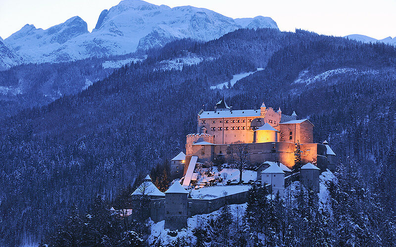 Ein großes, in der Abenddämmerung beleuchtetes Schloss steht auf einem verschneiten, bewaldeten Hügel, mit hohen, schneebedeckten Bergen im Hintergrund.