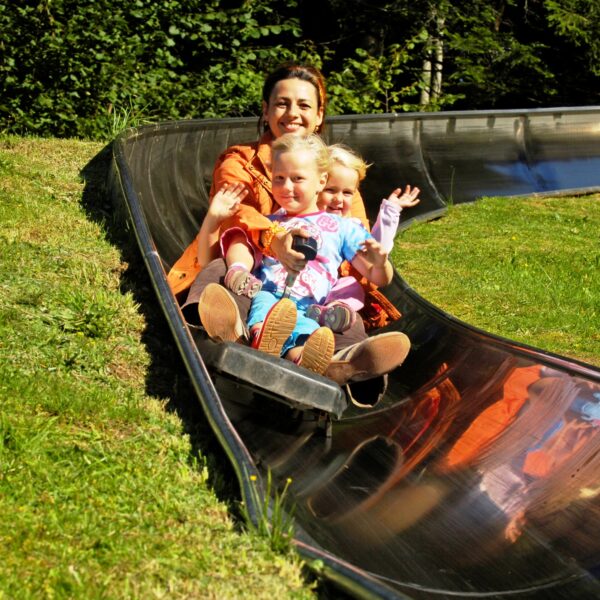 Ein Erwachsener und zwei Kinder fahren gemeinsam auf einer Metallrutsche im Freien, umgeben von grünem Gras und Bäumen.
