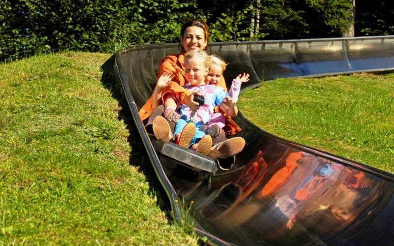 Ein Erwachsener und zwei Kinder fahren gemeinsam auf einer Metallrutsche im Freien, umgeben von grünem Gras und Bäumen.