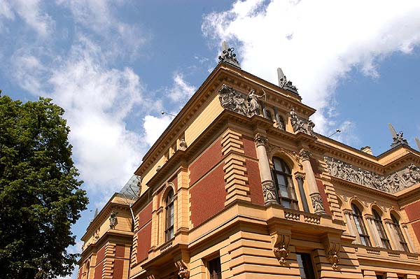 Reich verziertes historisches Gebäude mit Akzenten aus rotem Backstein und Stein, dekorativen Skulpturen, Bogenfenstern und einem bewölkten blauen Himmel im Hintergrund.