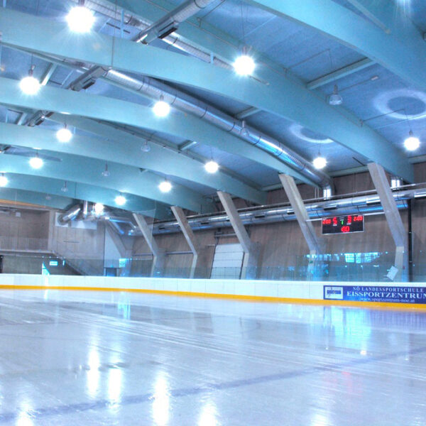 Eislaufhalle mit glattem, reflektierendem Eis, leeren Tribünen, Oberlicht und einer digitalen Anzeigetafel, die die Spielstände an der hinteren Wand anzeigt.