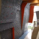 Indoor-Kletterwand mit verschiedenfarbigen Griffen und Sicherheitsmatten am Fuß, die sich neben einer Treppe in einer modernen Anlage befindet.