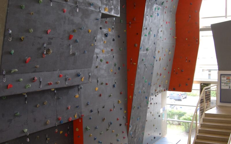 Indoor-Kletterwand mit verschiedenfarbigen Griffen und Sicherheitsmatten am Fuß, die sich neben einer Treppe in einer modernen Anlage befindet.
