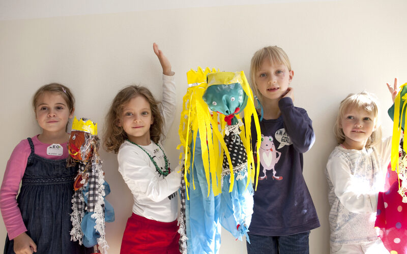 Vier kleine Kinder stehen drinnen und halten bunte, handgefertigte Kunstwerke mit Luftschlangen und Dekorationen in die Höhe und posieren vor einer schlichten weißen Wand.