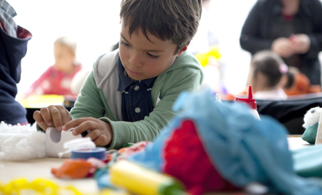 Ein kleiner Junge sitzt an einem Tisch und arbeitet an einem Bastelprojekt, umgeben von bunten Materialien und anderen Kindern im Hintergrund.